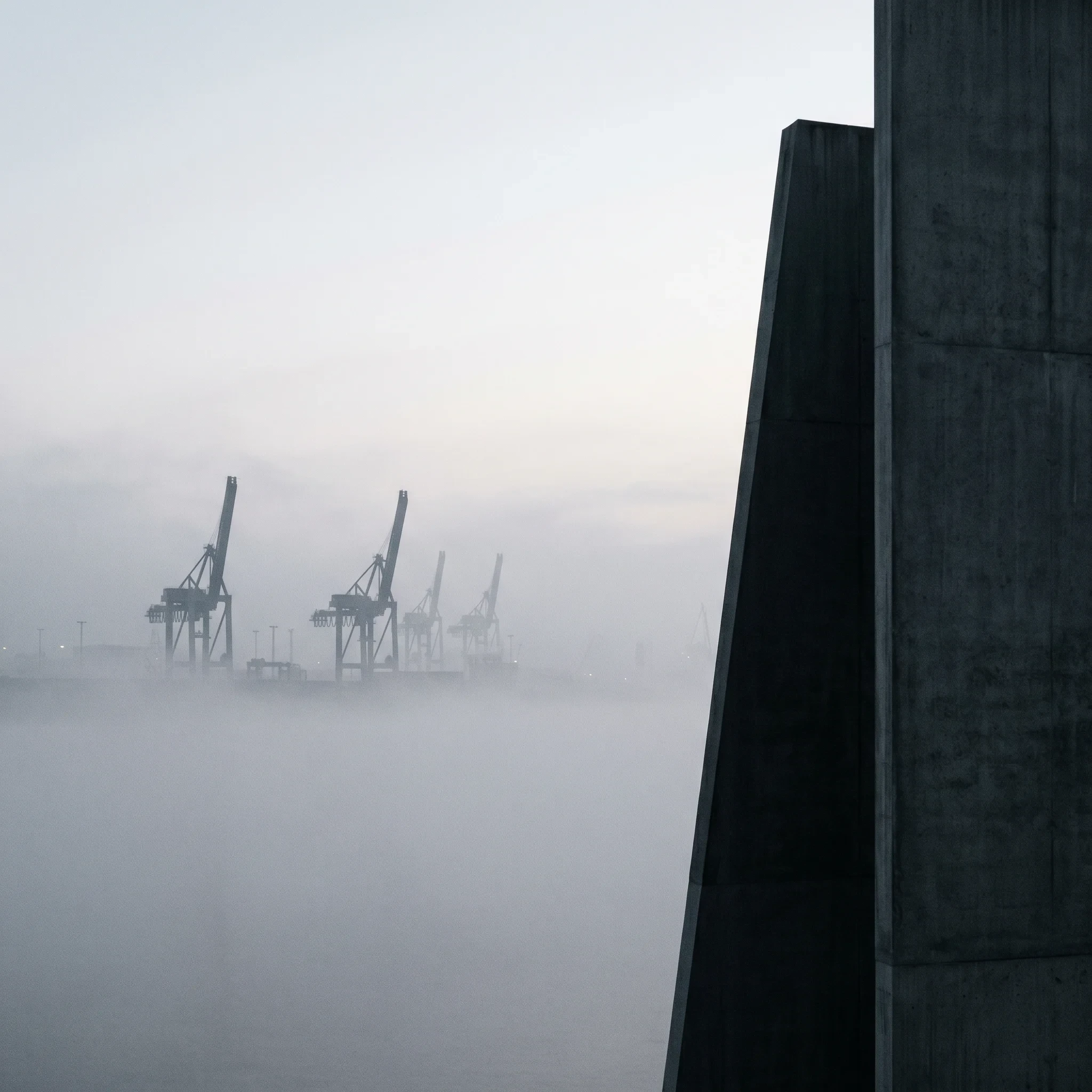 Foggy harbor cranes beside a dark architectural edge